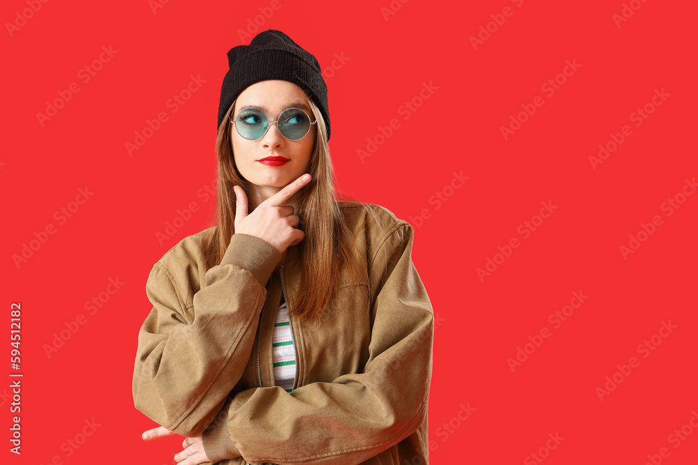 Thoughtful young woman in sunglasses pointing at something on red background