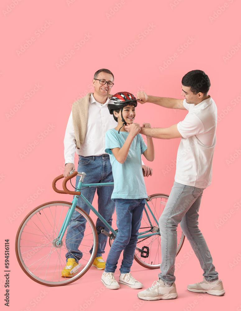 Young man putting bike helmet onto his little son and father on pink background