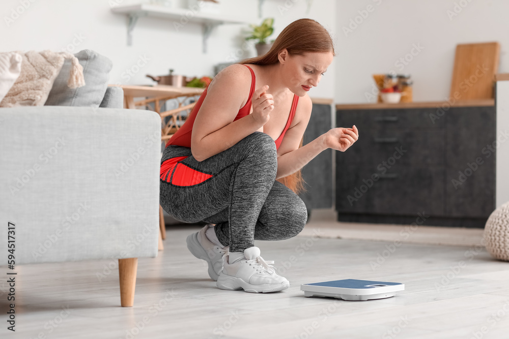 Upset young overweight woman measuring her weight on scales in kitchen