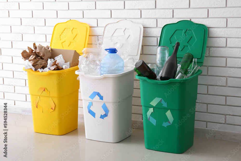 Trash bins with recycling symbol and different garbage near white brick wall