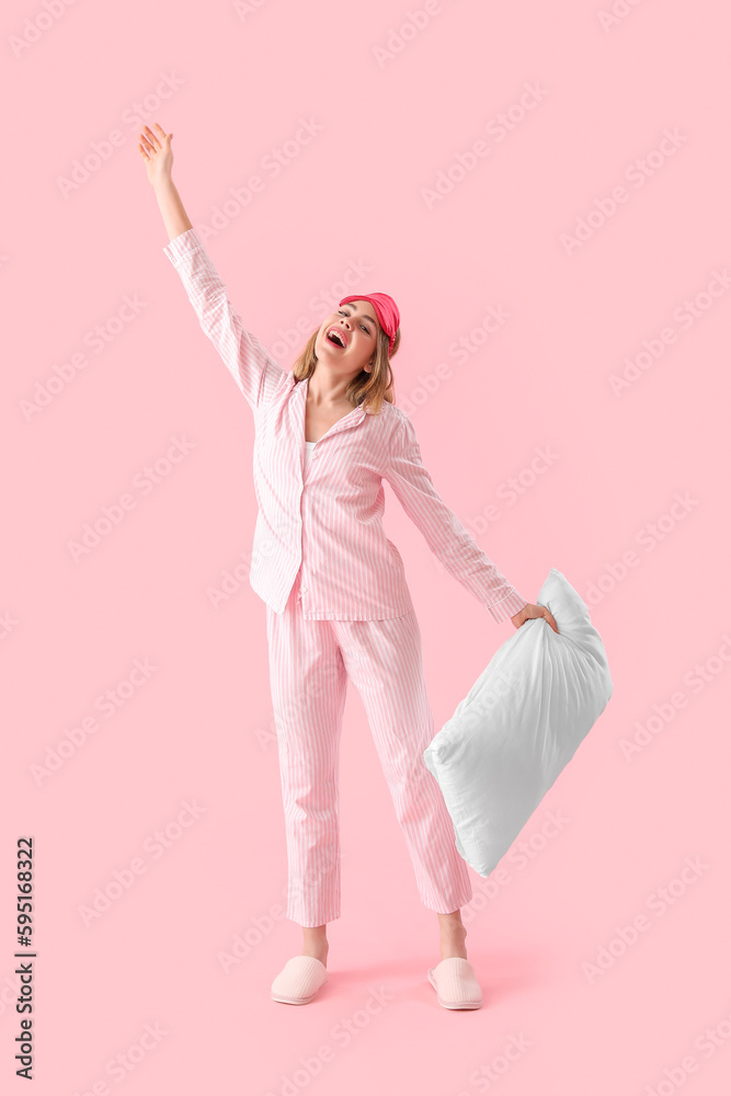 Happy young woman in pajamas with pillow on pink background