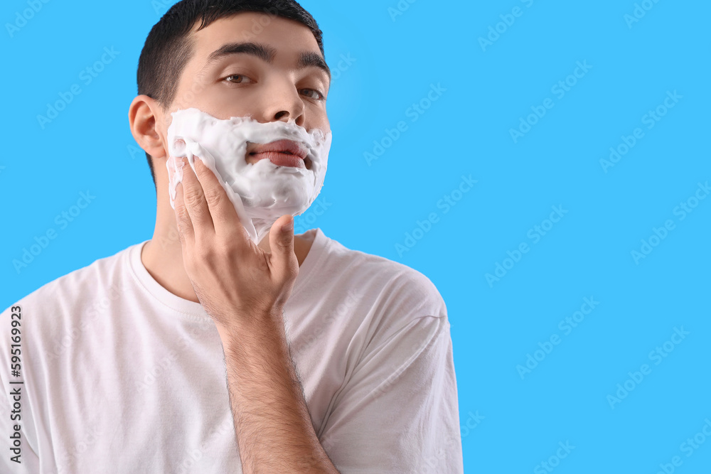 Young man with shaving foam on face against light blue background