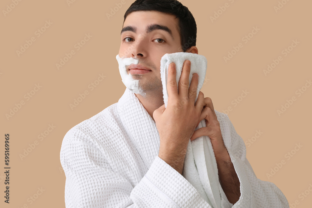 Young man wiping shaving foam from face against beige background