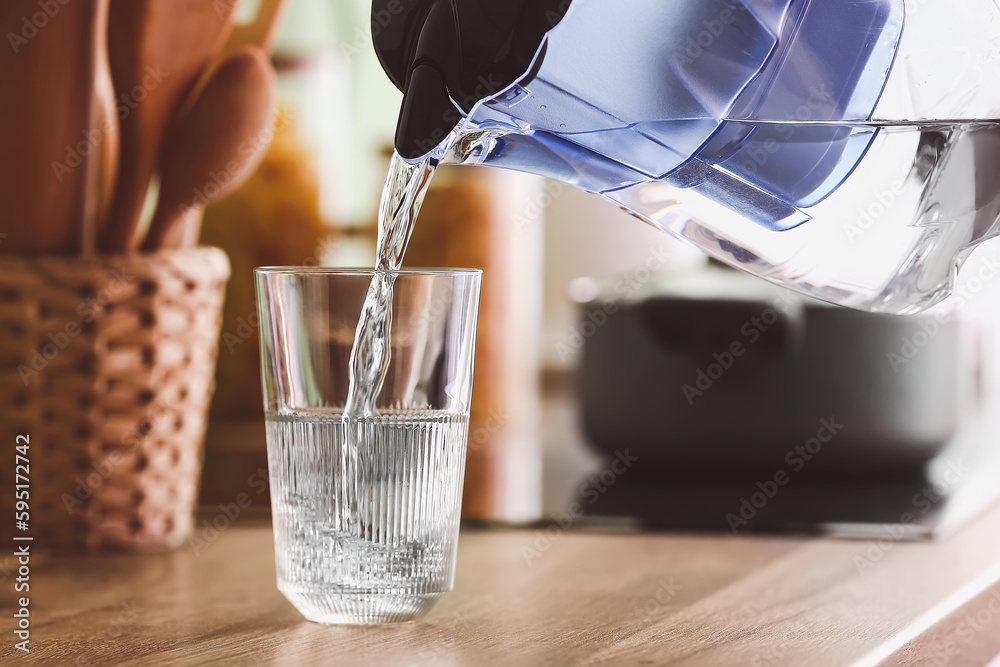 Pouring of water from modern filter jug into glass on kitchen counter