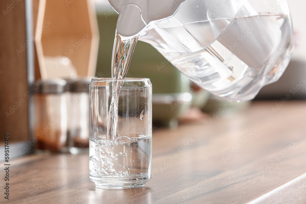 Pouring of water from modern filter jug into glass on kitchen counter