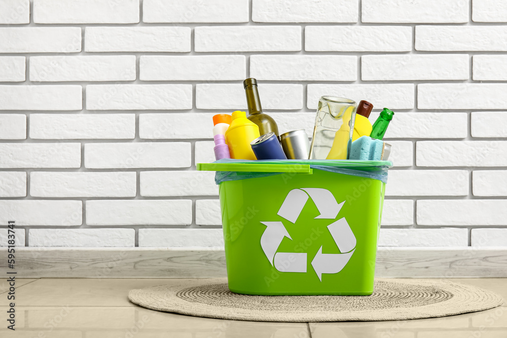 Trash bin with different garbage on rug near white brick wall