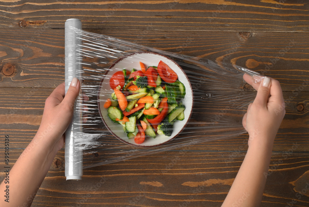 Female hands wrapping bowl with salad by stretch wrap on wooden background