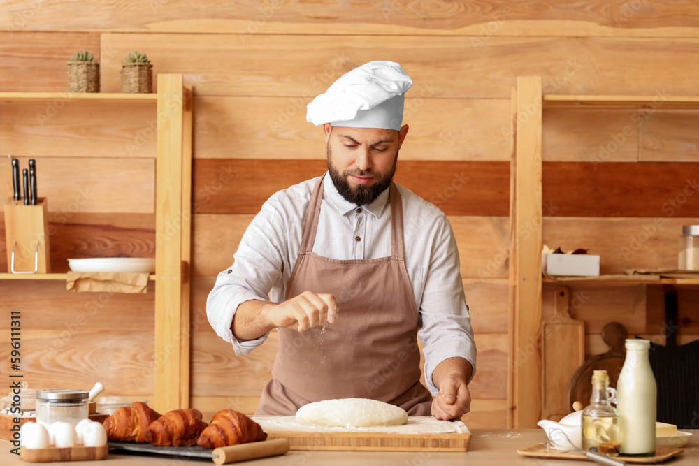 Male baker sprinkling dough with flour at table in kitchen