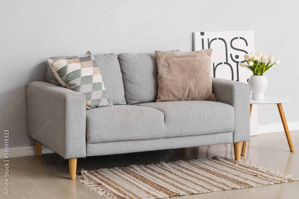 Cozy sofa with cushions and tulip flowers in vase on table near grey wall