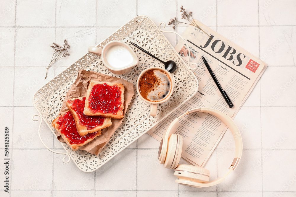 Tray with cup of coffee, milk, toasts, headphones, newspaper and flowers on white tile table