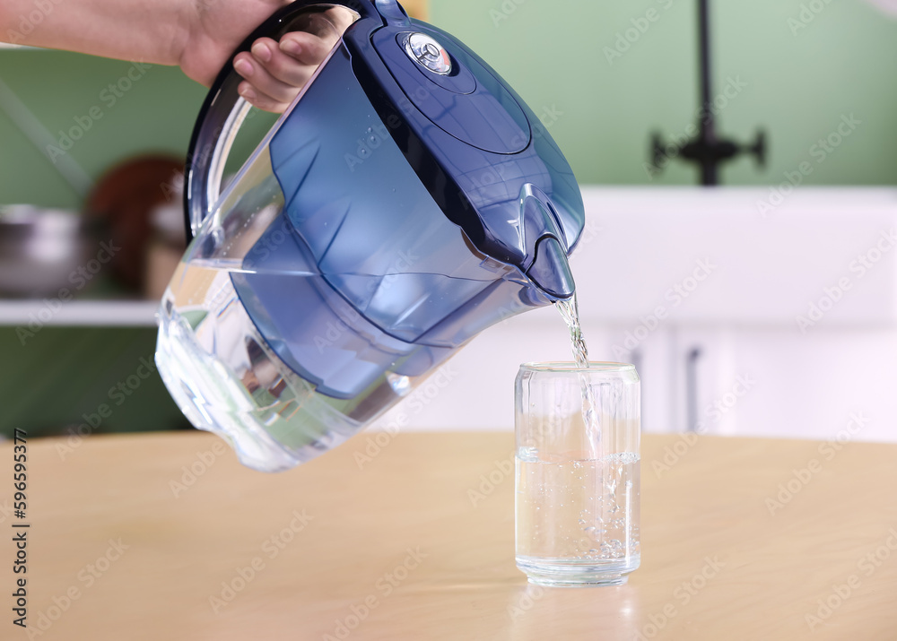 Woman pouring water from modern filter jug into glass on wooden table in kitchen