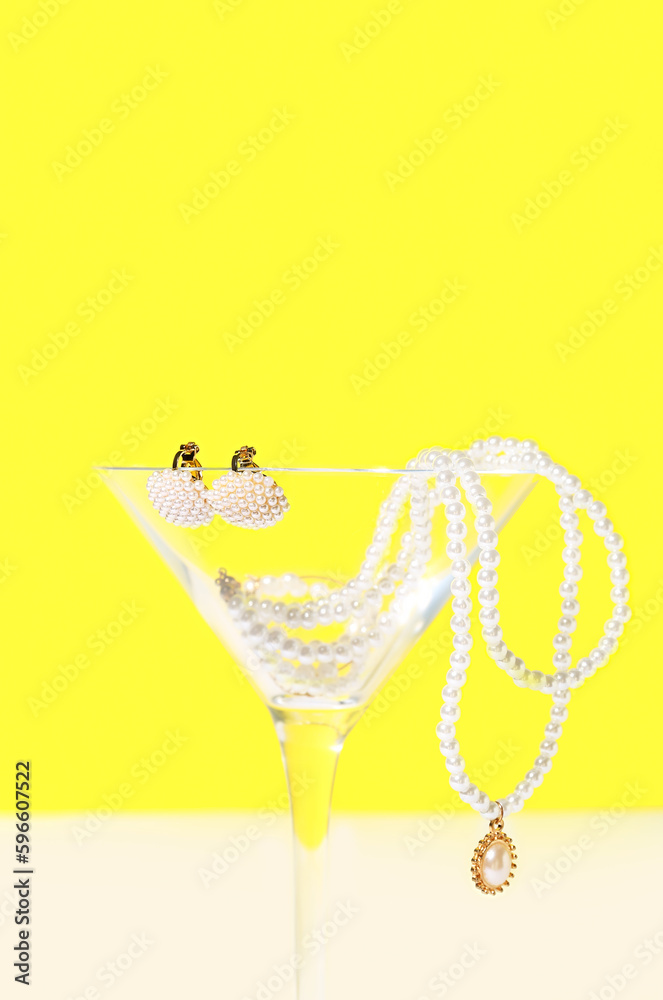 Glass with pearl jewelry on table against yellow background