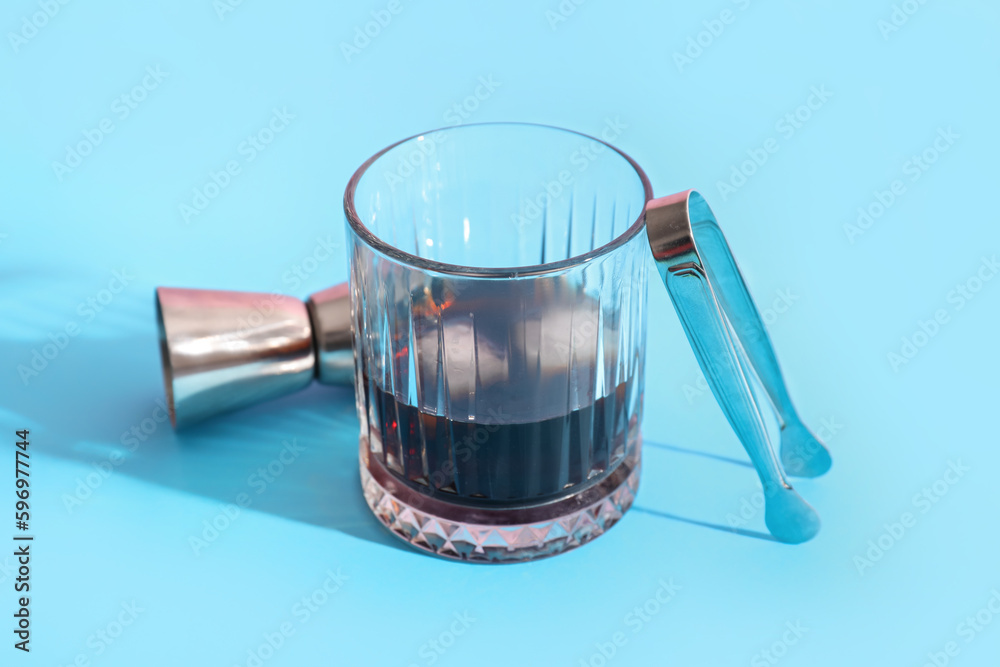 Glass of rum with ice cubes, tongs and measure cup on blue background, closeup