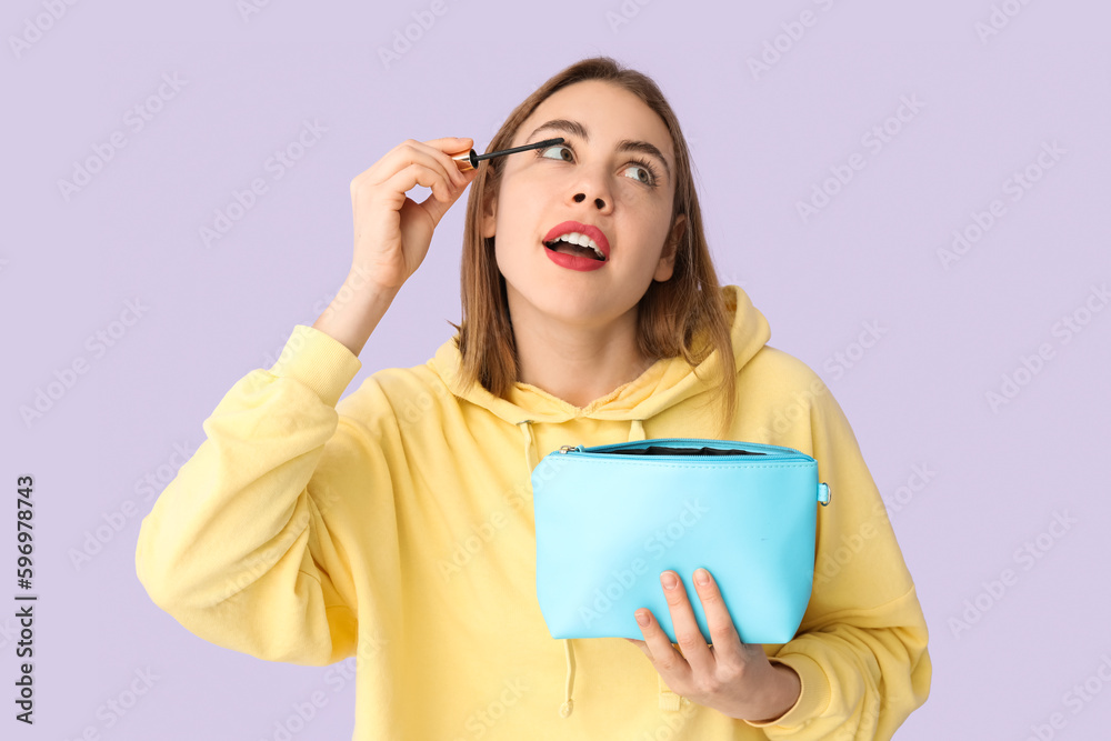 Young woman with cosmetic bag applying mascara on lilac background