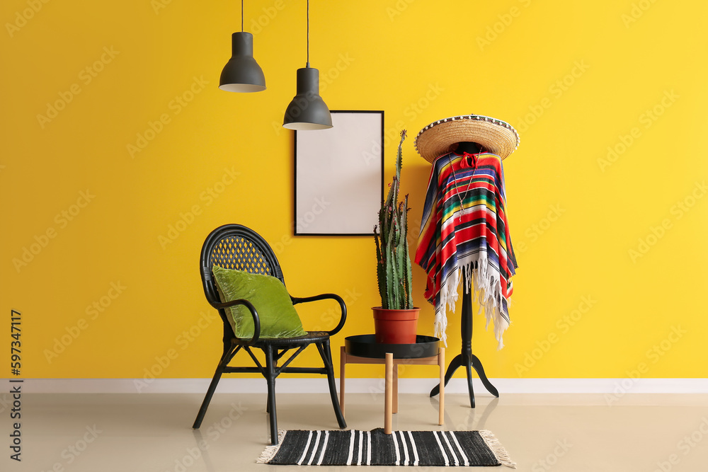 Rack with Mexican clothes, armchair and cactus on table near yellow wall