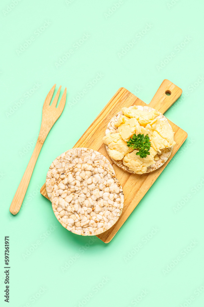 Wooden board with tasty scrambled eggs and rice crackers on green background