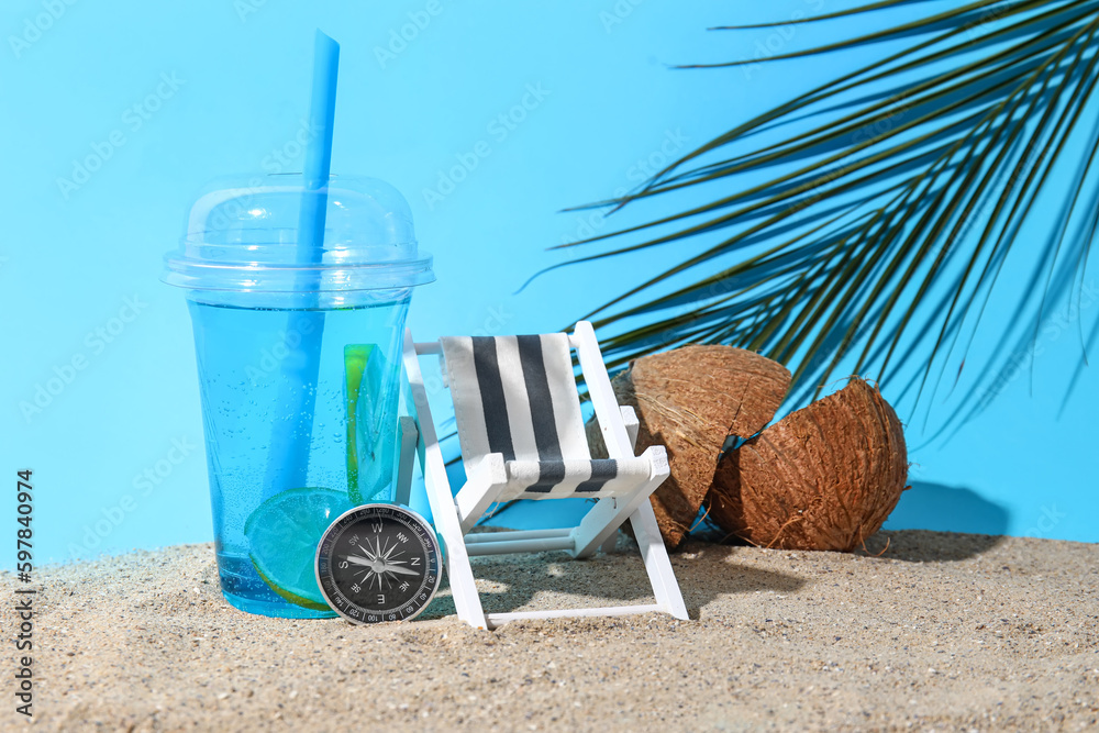 Composition with miniature deck chair, drink, compass and coconut in sand on blue background