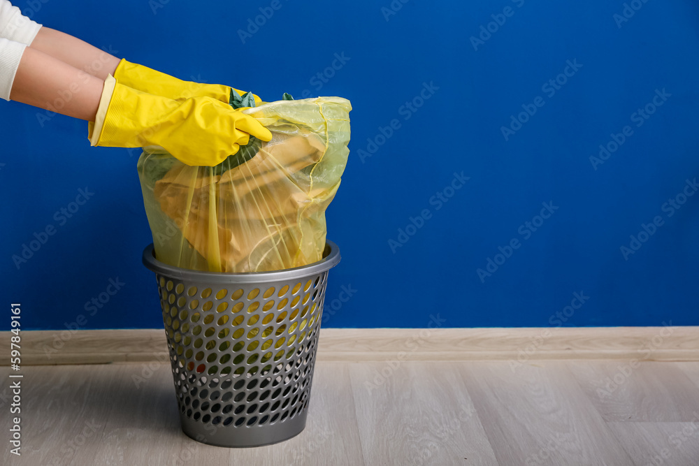 Female hands in rubber gloves pulling full garbage bag from trash bin near blue wall