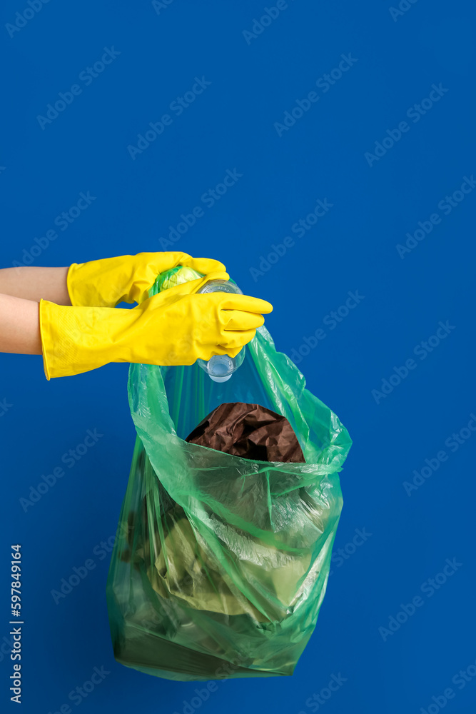 Female hands in rubber gloves putting plastic bottle into garbage bag on blue background