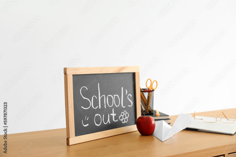 Chalkboard with text SCHOOLS OUT, apple, paper plane and stationery on table near light wall