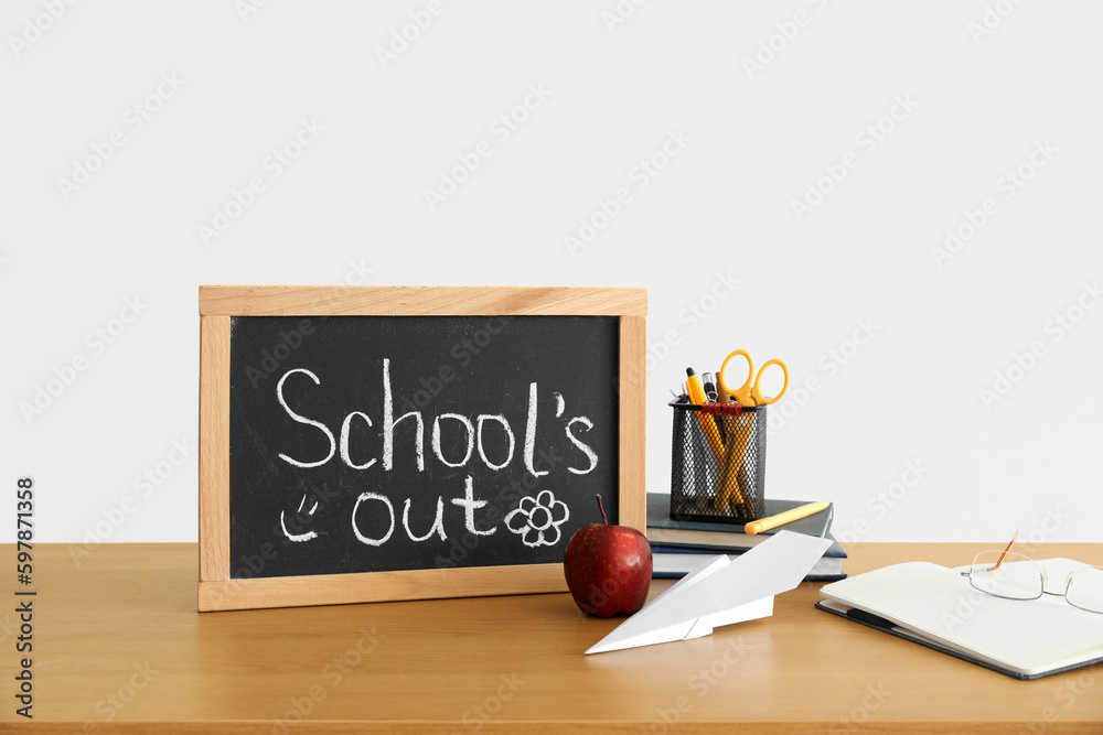 Chalkboard with text SCHOOLS OUT, apple, paper plane and stationery on table near light wall