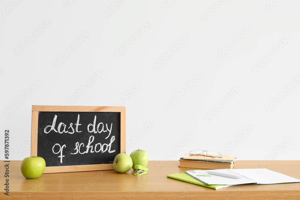 Chalkboard with text LAST DAY OF SCHOOL, apples and notebooks on table near light wall