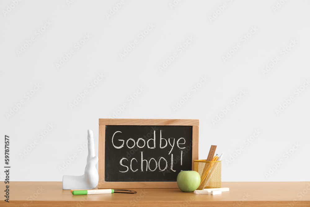 Chalkboard with text GOODBYE SCHOOL, apple and stationery on table near light wall