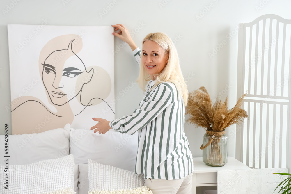 Mature woman hanging painting on light wall in bedroom