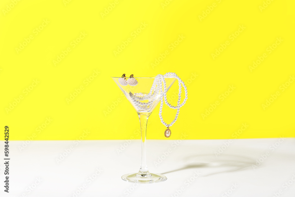 Glass with pearl jewelry on table against yellow background