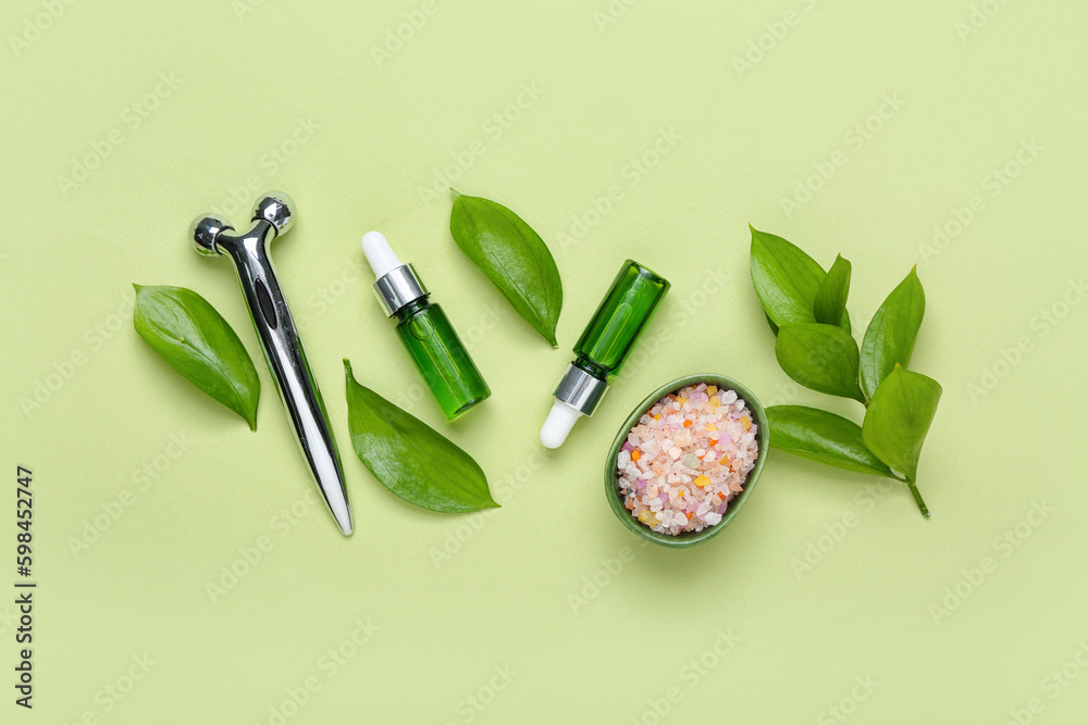 Bottles of cosmetic oil with plant leaves, massage roller and sea salt on light green background