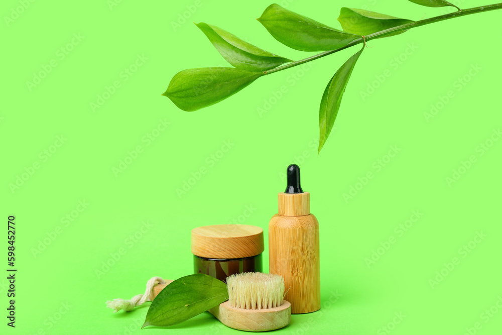 Bottles of cosmetic oil with plant twig, jar of cream and massage brush on light green background