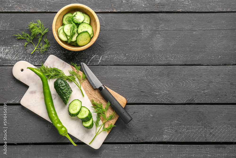 Board and bowl with fresh cut cucumber on black wooden background