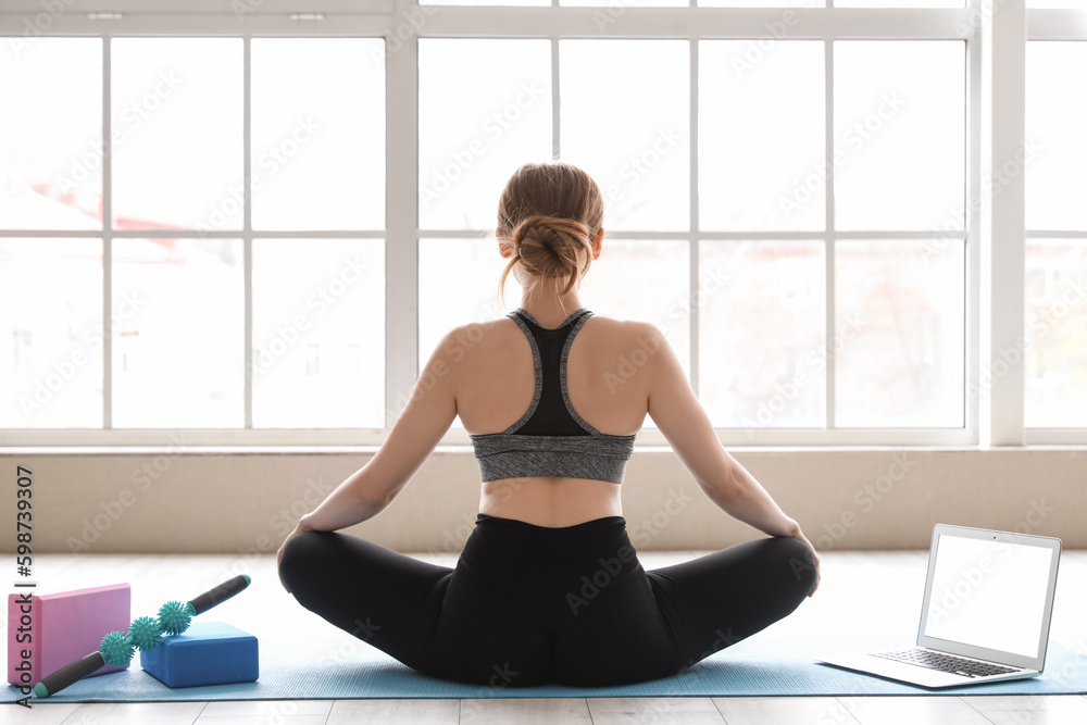 Sporty young woman with laptop doing yoga in gym, back view