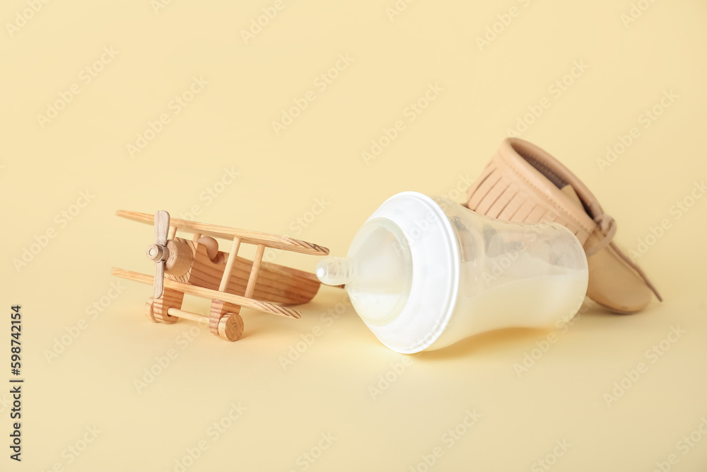 Bottle of milk for baby with toy and booties on pale yellow background