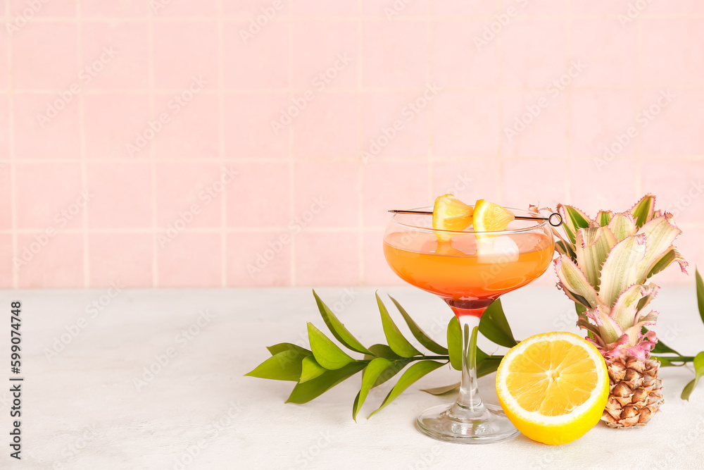 Glass of cocktail with lemon, mini pineapple and plant branch on table near pink tile wall