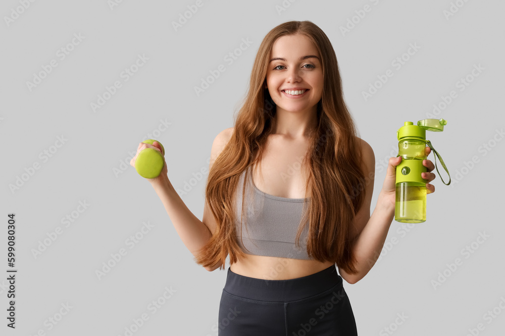 Sporty young woman with bottle of water and dumbbell on grey background