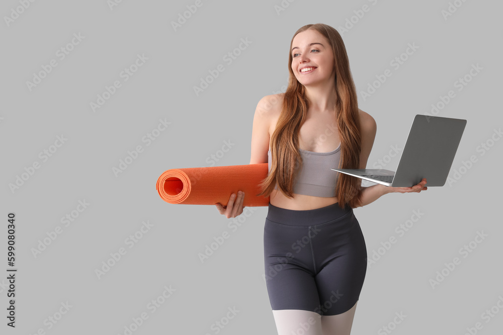 Sporty young woman with yoga mat and laptop on grey background