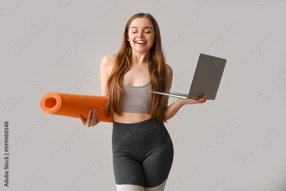 Sporty young woman with yoga mat and laptop on grey background