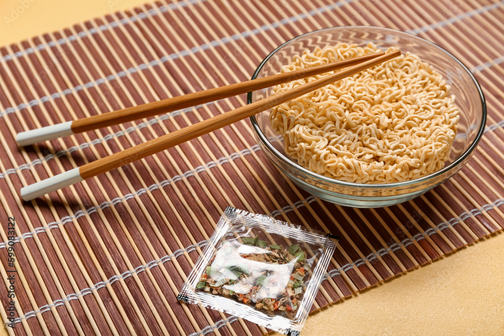 Raw instant noodles in bowl with chopsticks, seasoning and bamboo mat on yellow background