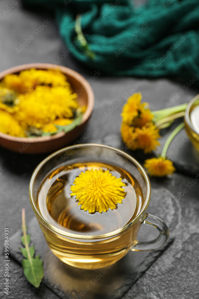 Glass cup of healthy dandelion tea and bowl with flowers on black background