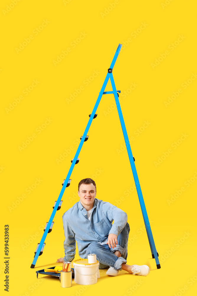 Young man with ladder and cans of paint sitting on yellow background