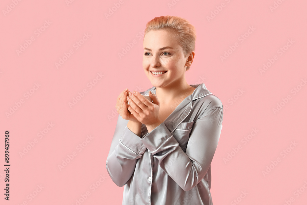 Young woman in pajamas with cup of coffee on pink background