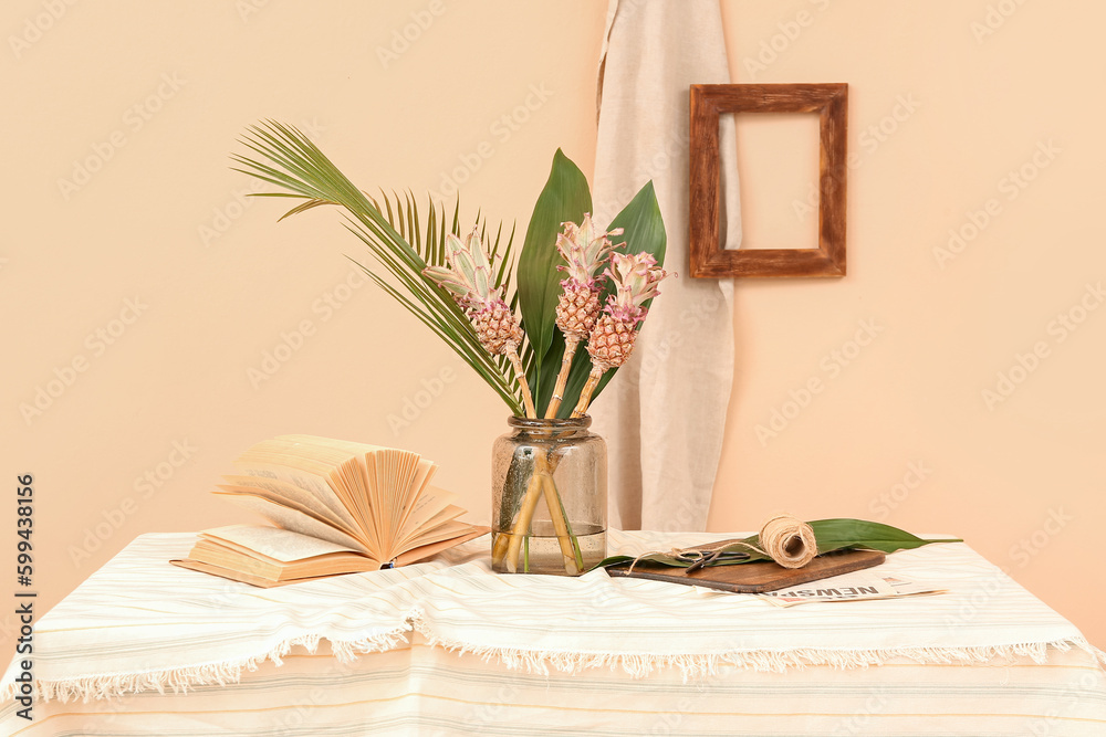 Vase with decorative pineapples, palm leaves, book and rope on table near beige wall