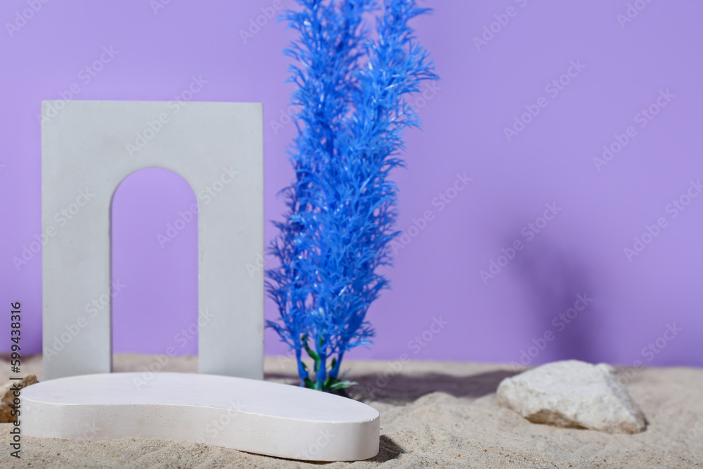 Decorative plaster podiums, seaweed and stones in sand on lilac background