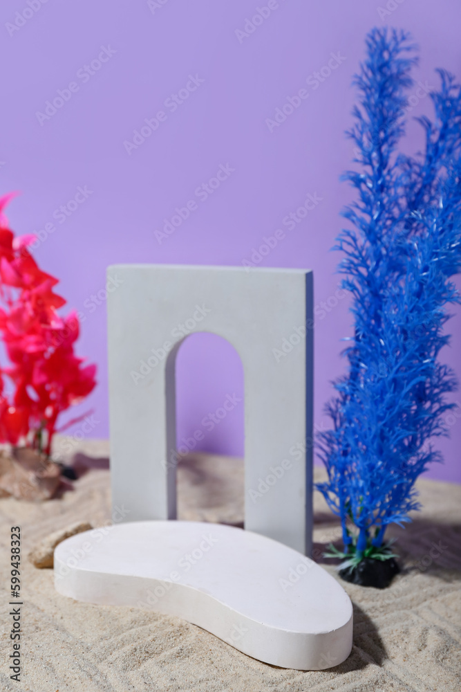 Decorative plaster podiums, seaweed and stones in sand on lilac background