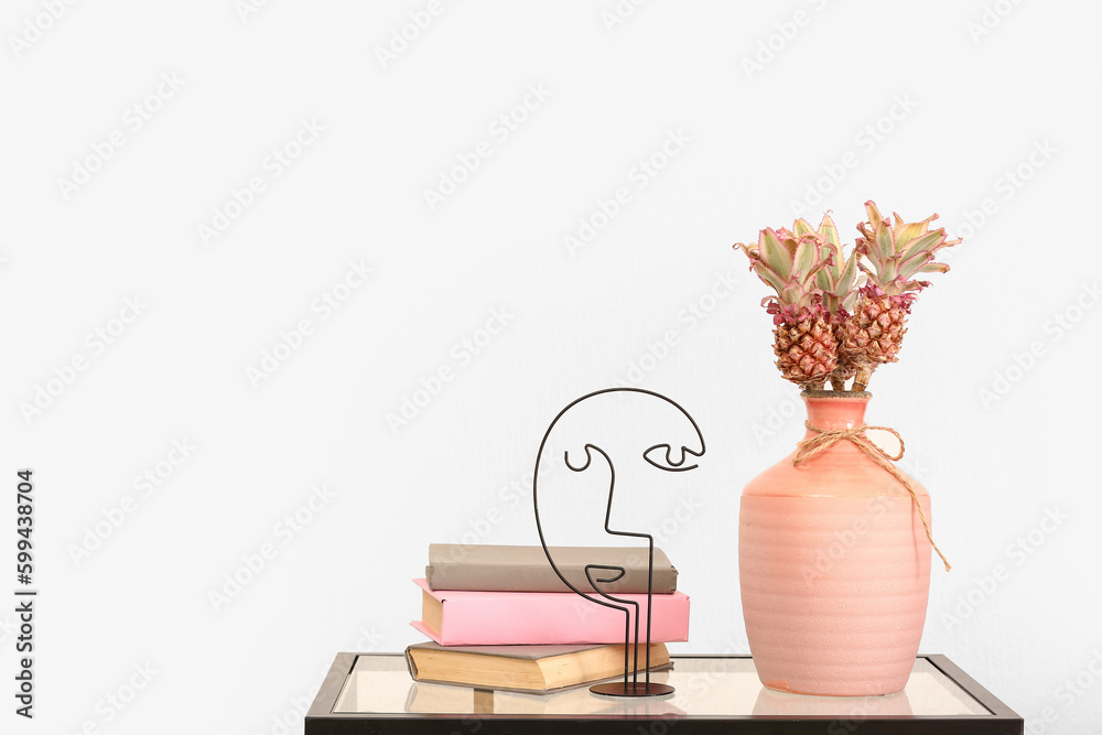 Vase with baby pineapples, books and decor on table near light wall