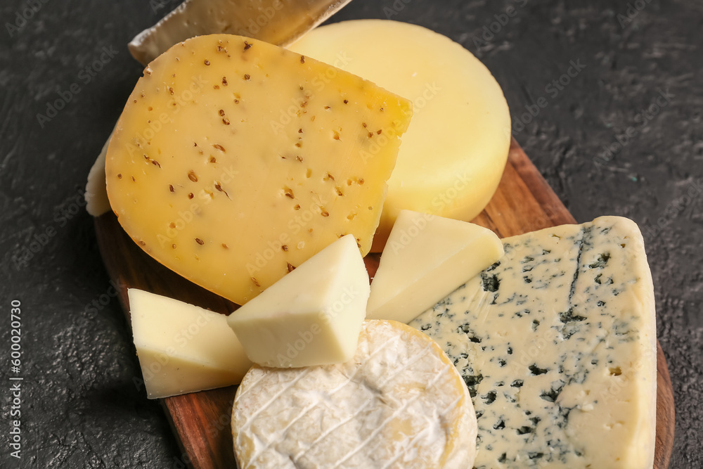 Wooden board with different types of tasty cheese on dark background, closeup