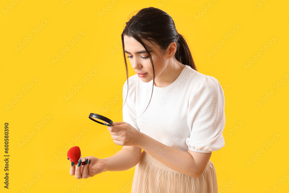 Young woman with magnifier and engagement ring on yellow background