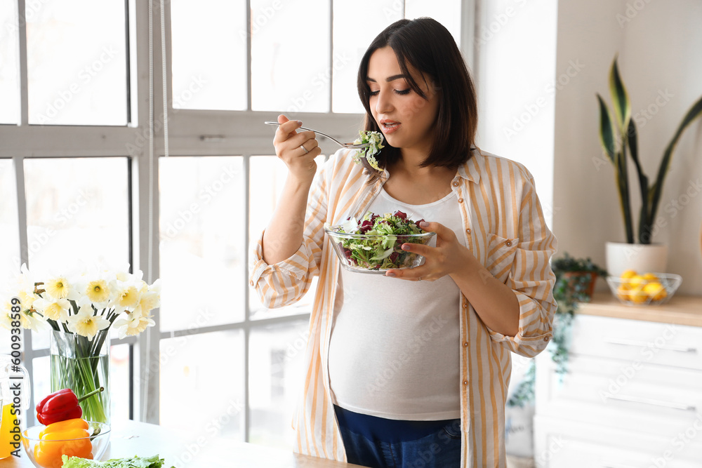 Young pregnant woman eating vegetable salad in kitchen