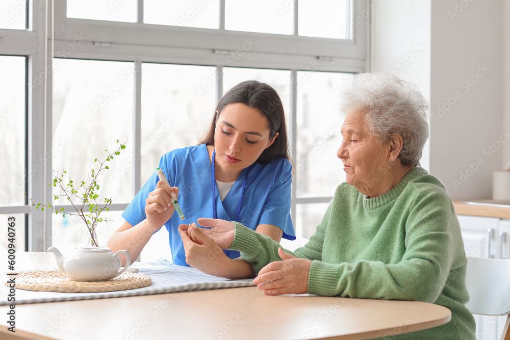 Young caregiver measuring sugar level of senior woman in kitchen